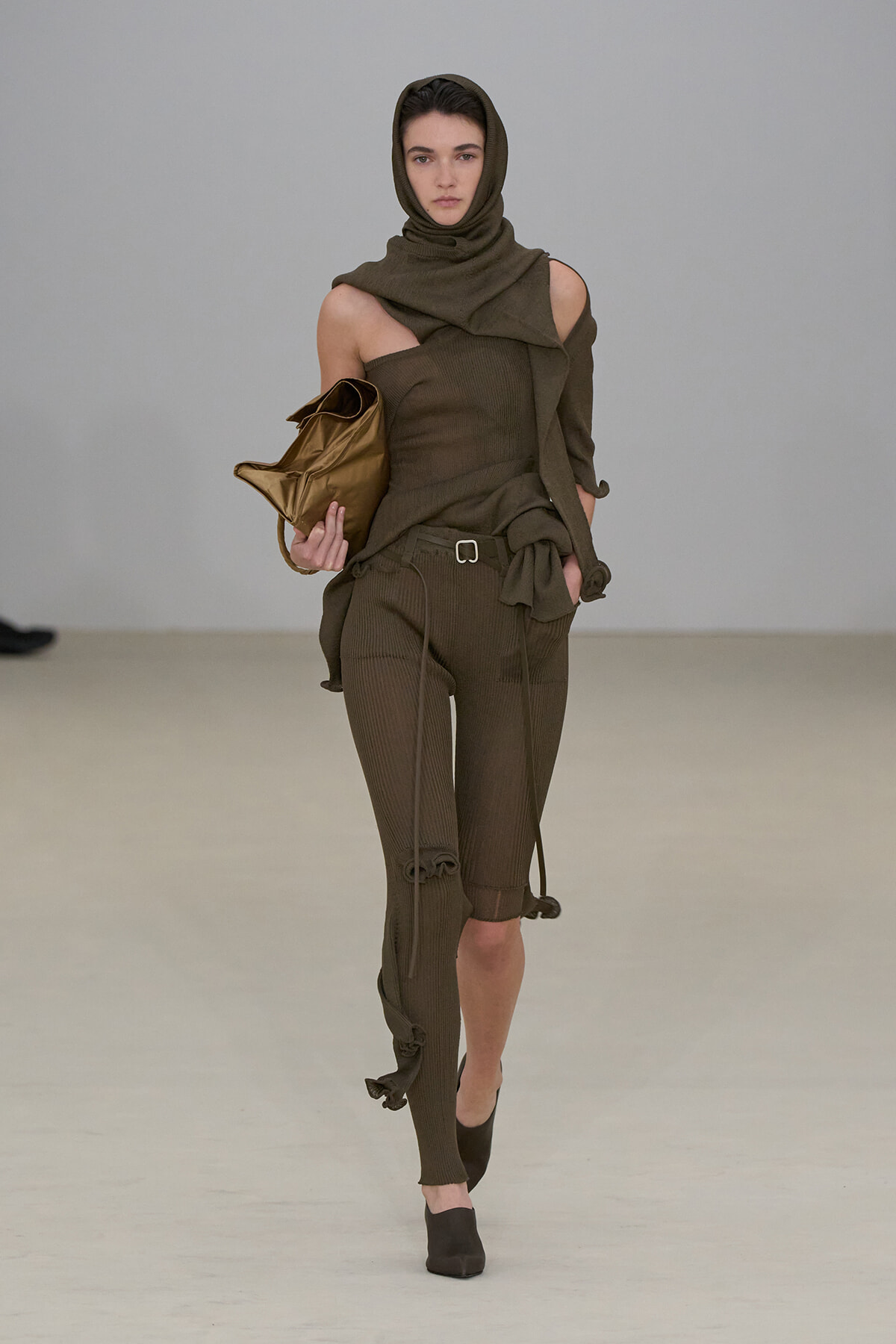 Model walking a runway in an olive-brown knit outfit with a hood, one-shoulder top, and belted pants, carrying a bronze clutch.