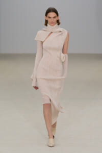 Model walks the runway in a pale beige ribbed knit dress with an asymmetrical neckline and scarf detail, paired with matching gloves and shoes.