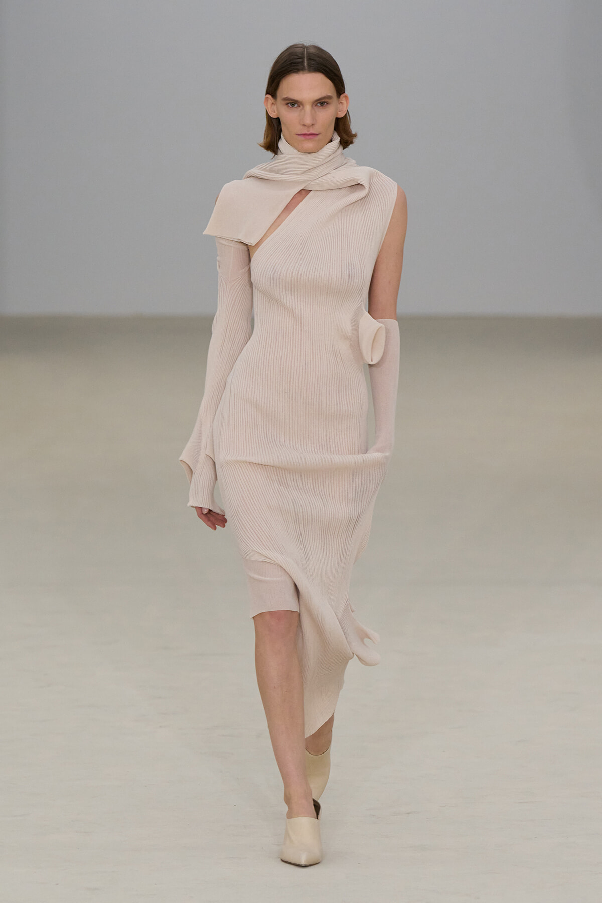 Model walks the runway in a pale beige ribbed knit dress with an asymmetrical neckline and scarf detail, paired with matching gloves and shoes.