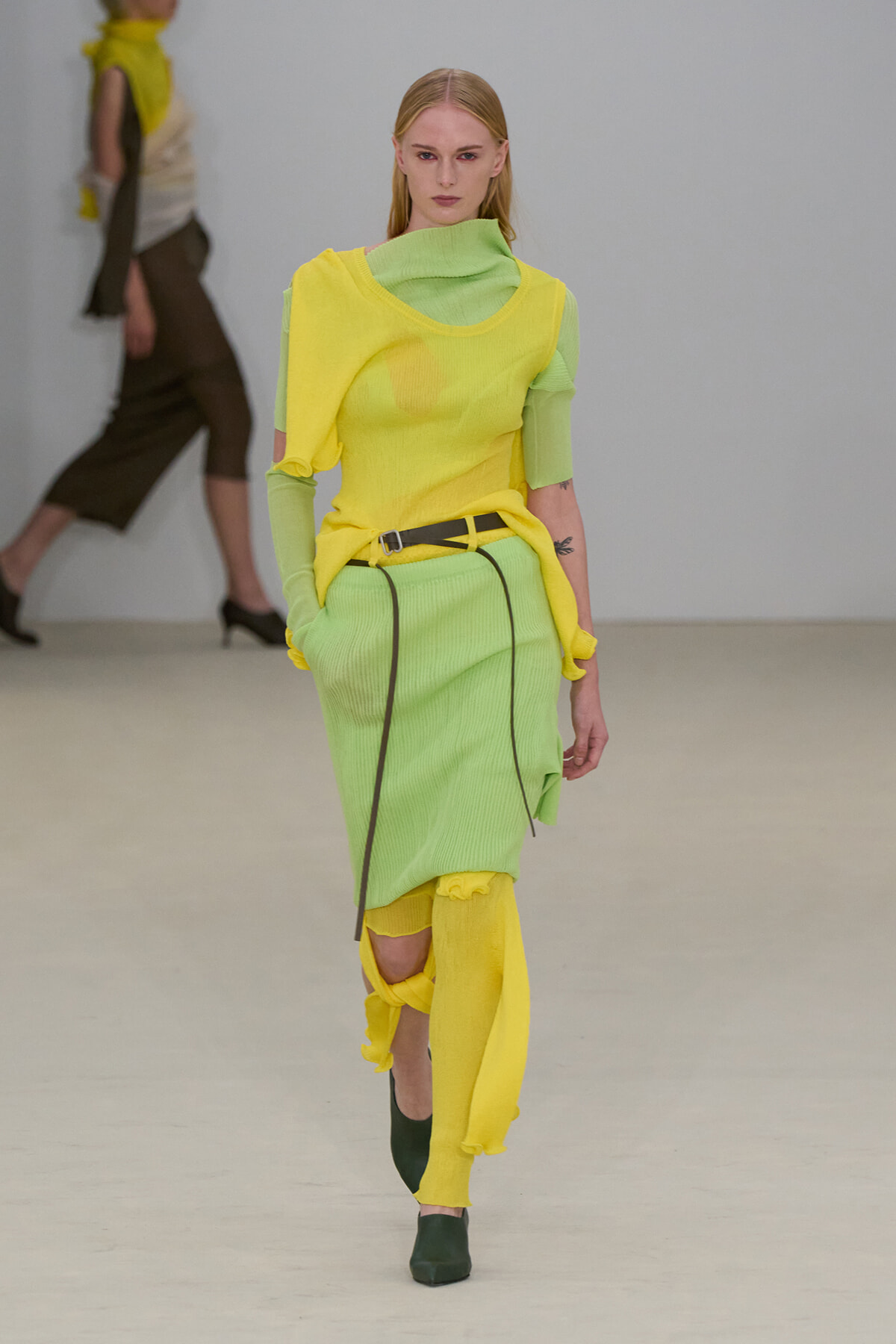 Model walking a fashion runway in a lime green knit dress layered with a bright yellow top and matching leg warmers, with a black belt and dark green boots.