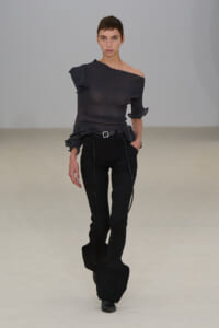 Model walks a runway in an all-black knit outfit: an off-the-shoulder top with a knot detail and high-waisted ribbed pants with a belt and long, sculpted cuffs at the ankles.