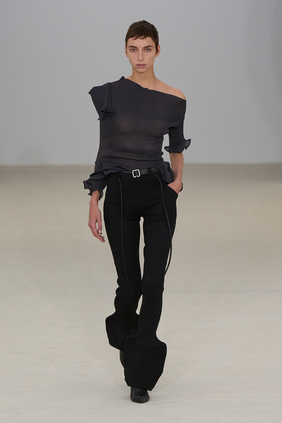 Model walks a runway in an all-black knit outfit: an off-the-shoulder top with a knot detail and high-waisted ribbed pants with a belt and long, sculpted cuffs at the ankles.