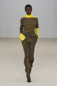 Model walking a fashion runway in an olive ribbed off-the-shoulder top paired with neon yellow turtleneck and cuffs, matching olive pants with belt and foldable hems.