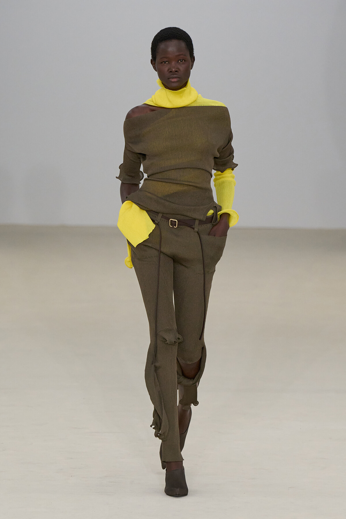 Model walking a fashion runway in an olive ribbed off-the-shoulder top paired with neon yellow turtleneck and cuffs, matching olive pants with belt and foldable hems.