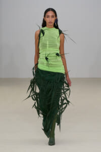 Model walks a runway in a neon lime sleeveless top paired with a dark green fringe skirt featuring long leaf-like strips suggesting plant motifs.
