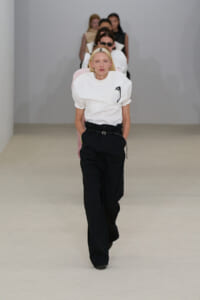 Model walking a fashion runway in a white puff-sleeve top and black wide-leg trousers with a belt; a line of seated/following models behind.
