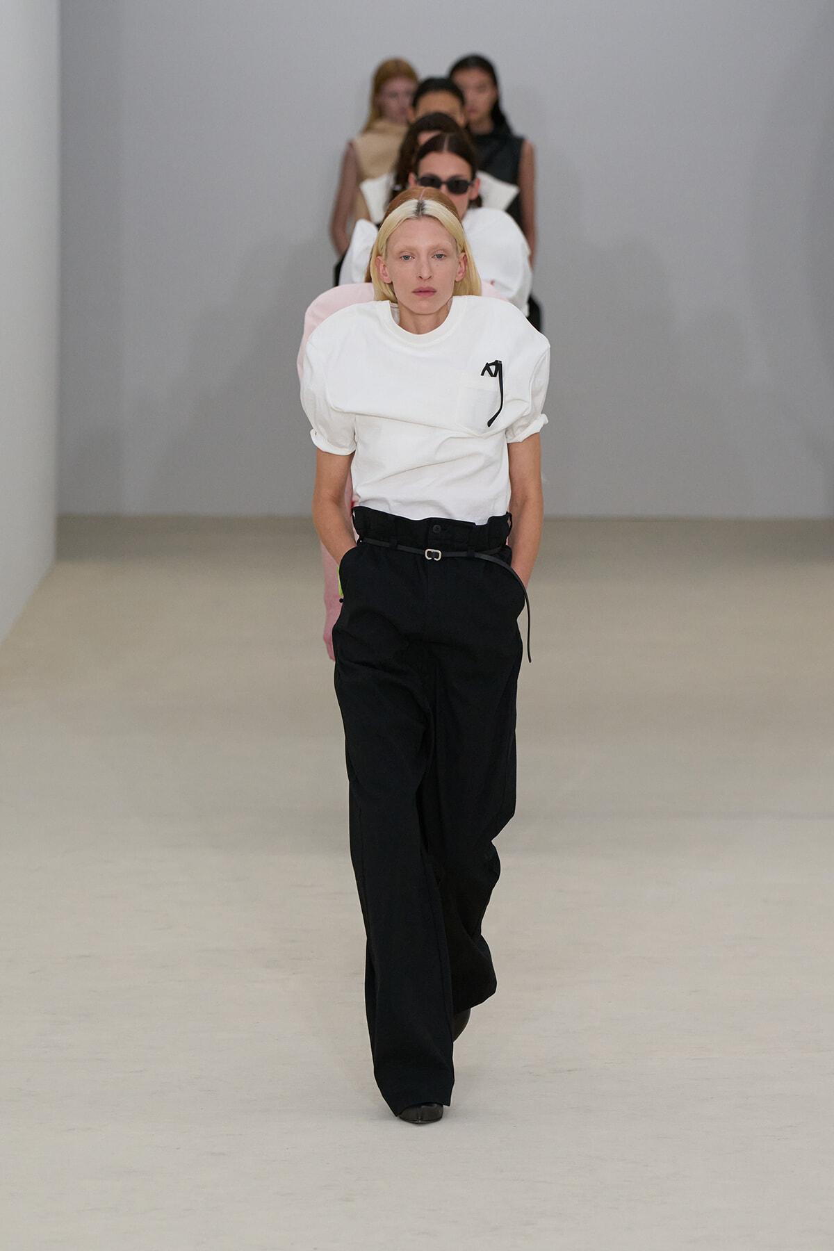 Model walking a fashion runway in a white puff-sleeve top and black wide-leg trousers with a belt; a line of seated/following models behind.