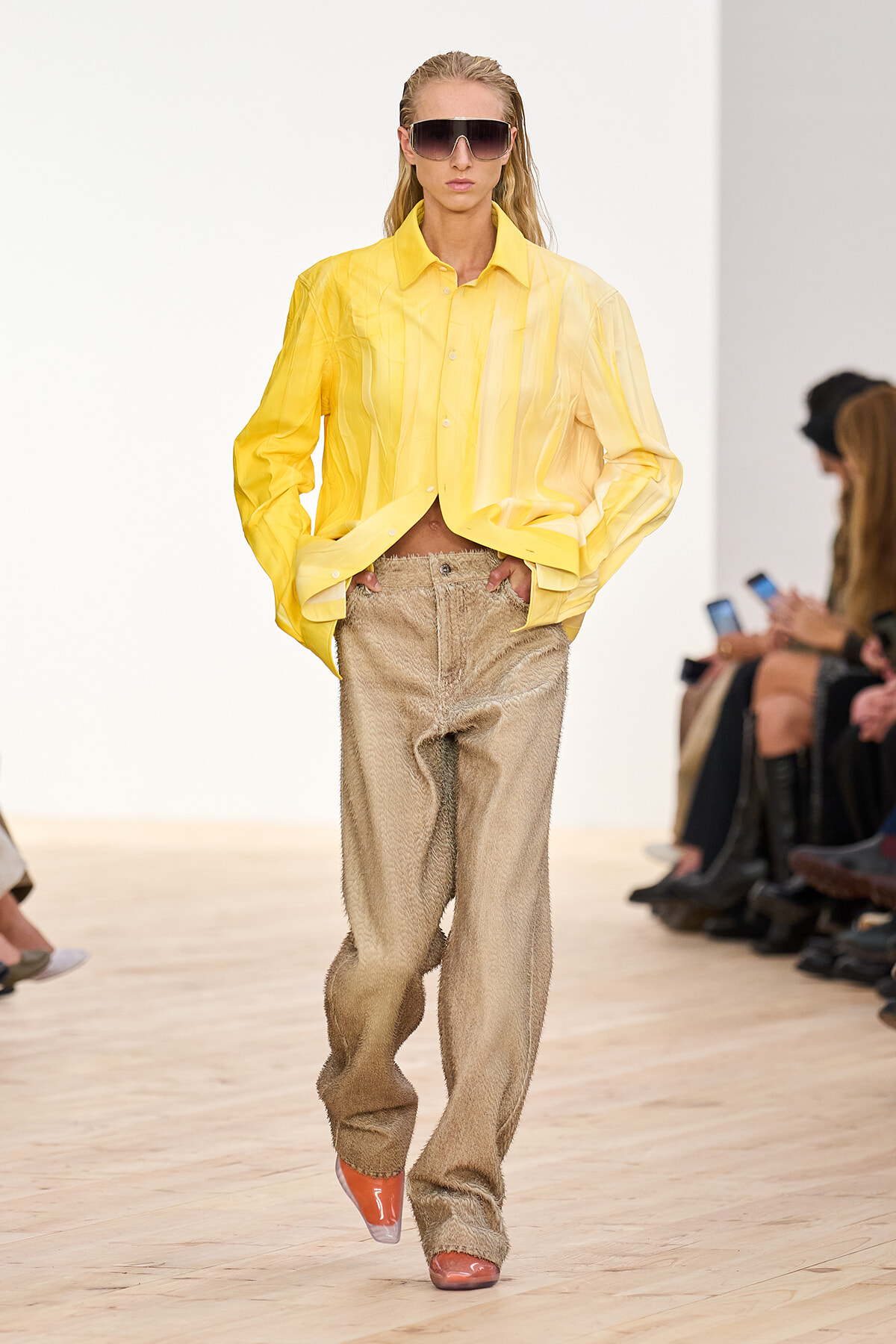 Runway model in an oversized yellow shirt with textured beige wide-leg pants and large sunglasses.