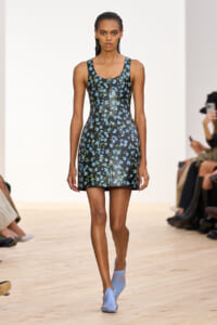 Model walking a fashion runway in a black mini dress with blue floral print and light blue shoes.