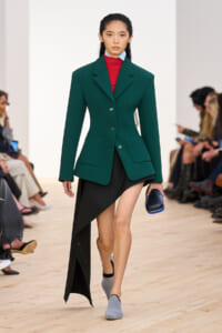 Model walks the runway in a dark green tailored blazer with broad shoulders over a red turtleneck and a black asymmetrical skirt, holding a blue clutch.