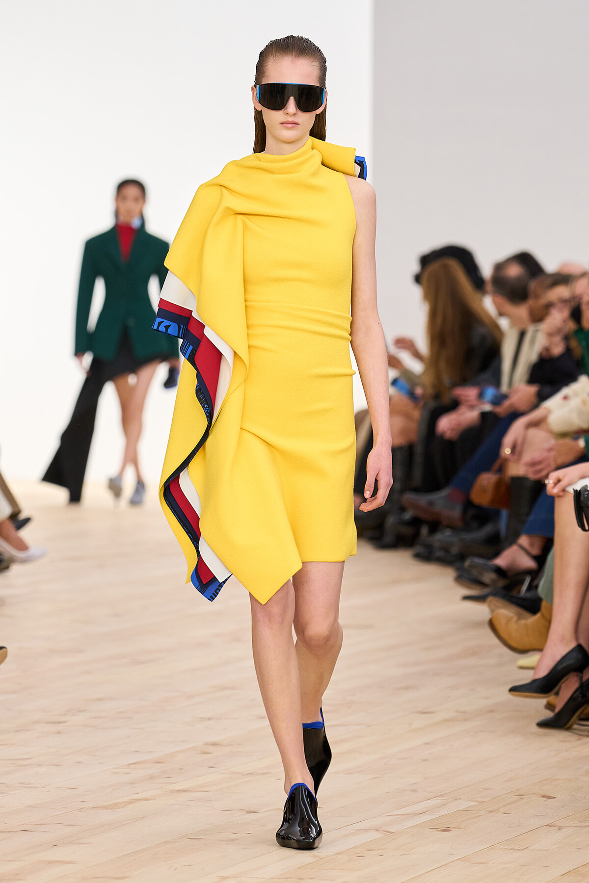 Model walks the runway in a bright yellow one-shoulder dress with a multicolor cape and oversized black sunglasses.
