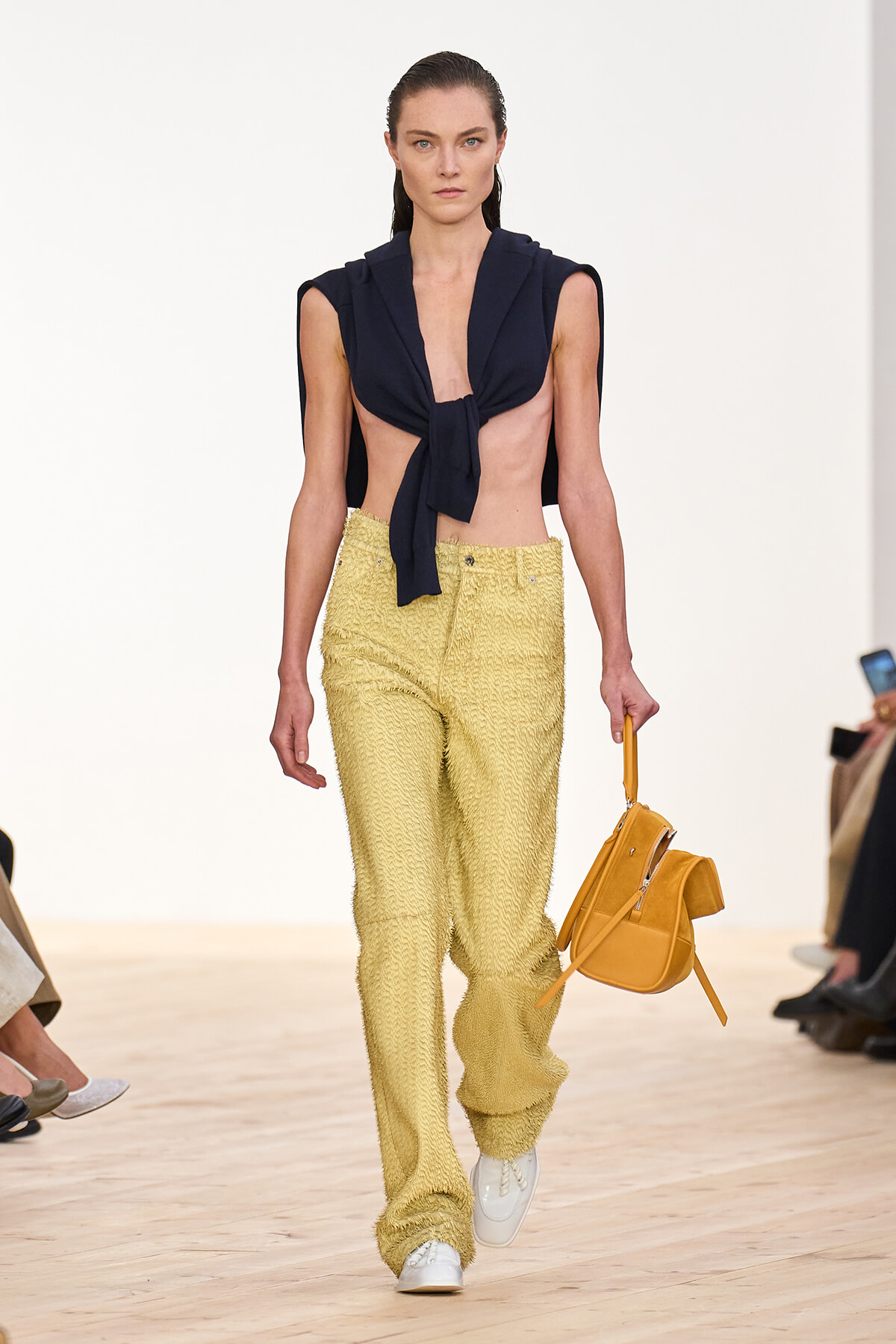 Model walking the runway in a navy sleeveless tie-front top and textured pale yellow pants, white sneakers, carrying a mustard handbag.