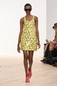 Female model walks a runway in a yellow floral mini dress with blue flowers, pink slip-on shoes, and oversized sunglasses.