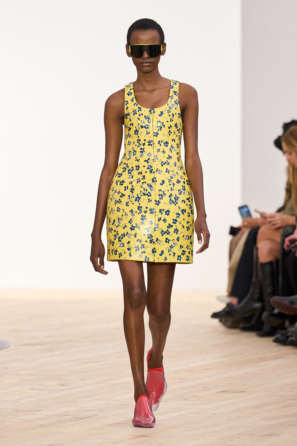 Female model walks a runway in a yellow floral mini dress with blue flowers, pink slip-on shoes, and oversized sunglasses.
