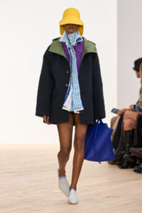 Model walking a runway in an oversized black coat with green collar, blue striped scarf, and a wide yellow hat, carrying a bright blue tote bag and wearing white slip-on shoes.