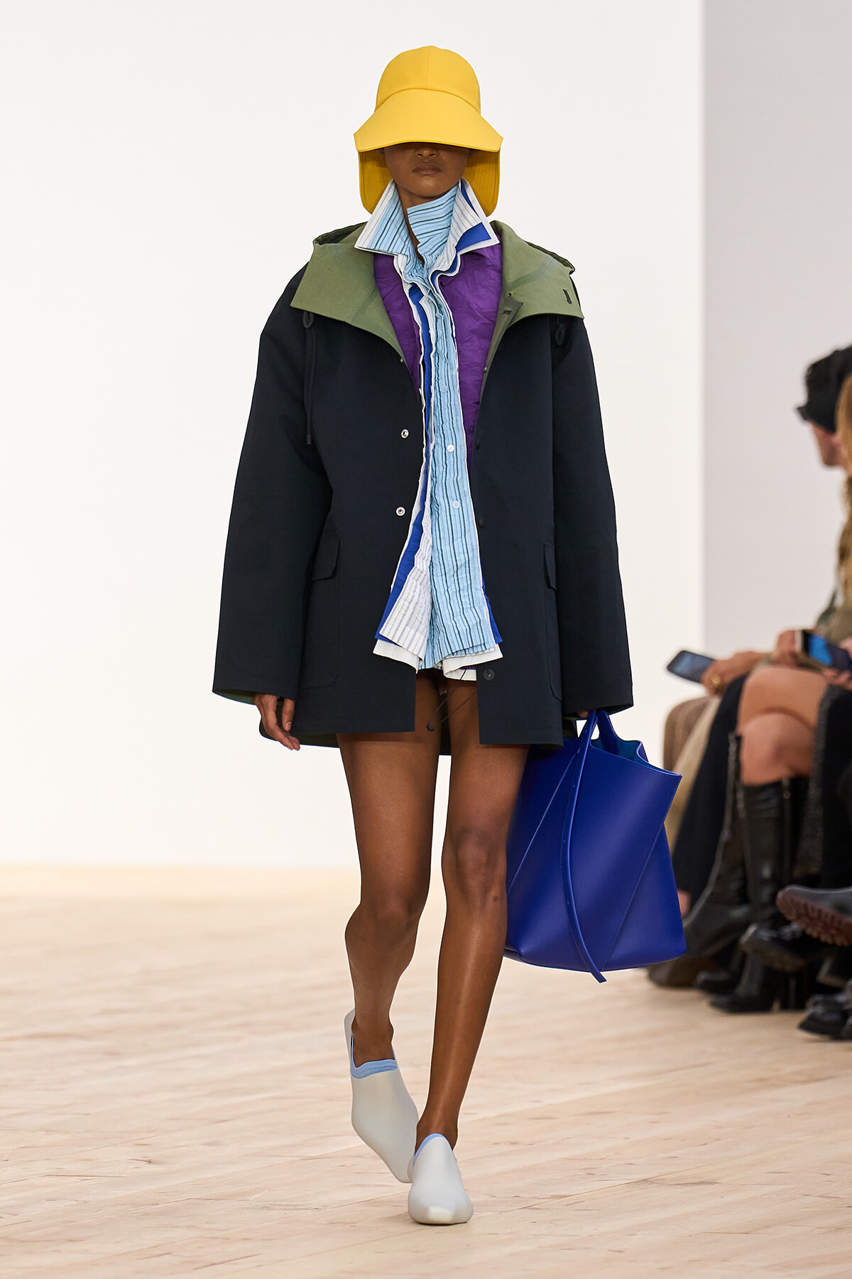Model walking a runway in an oversized black coat with green collar, blue striped scarf, and a wide yellow hat, carrying a bright blue tote bag and wearing white slip-on shoes.