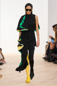 Runway model in a black sculptural dress with a green-yellow cape, paired with yellow and black tights and oversized sunglasses.