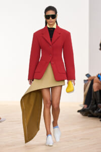 Model on a runway wearing a red structured blazer over a beige high-low dress, carrying a small yellow clutch and wearing futuristic dark sunglasses.