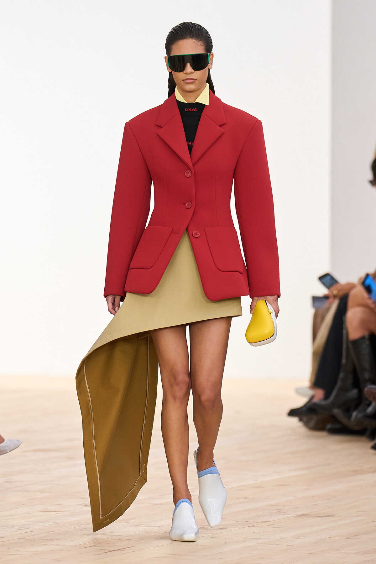 Model on a runway wearing a red structured blazer over a beige high-low dress, carrying a small yellow clutch and wearing futuristic dark sunglasses.