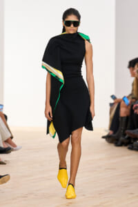 Model walking a runway in a black asymmetric dress with a multicolor cape, oversized sunglasses, and bright yellow mules.