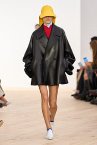 Model walking runway in an oversized black leather coat, bright yellow bucket hat, red top, and blue scarf, with white-and-blue shoes.