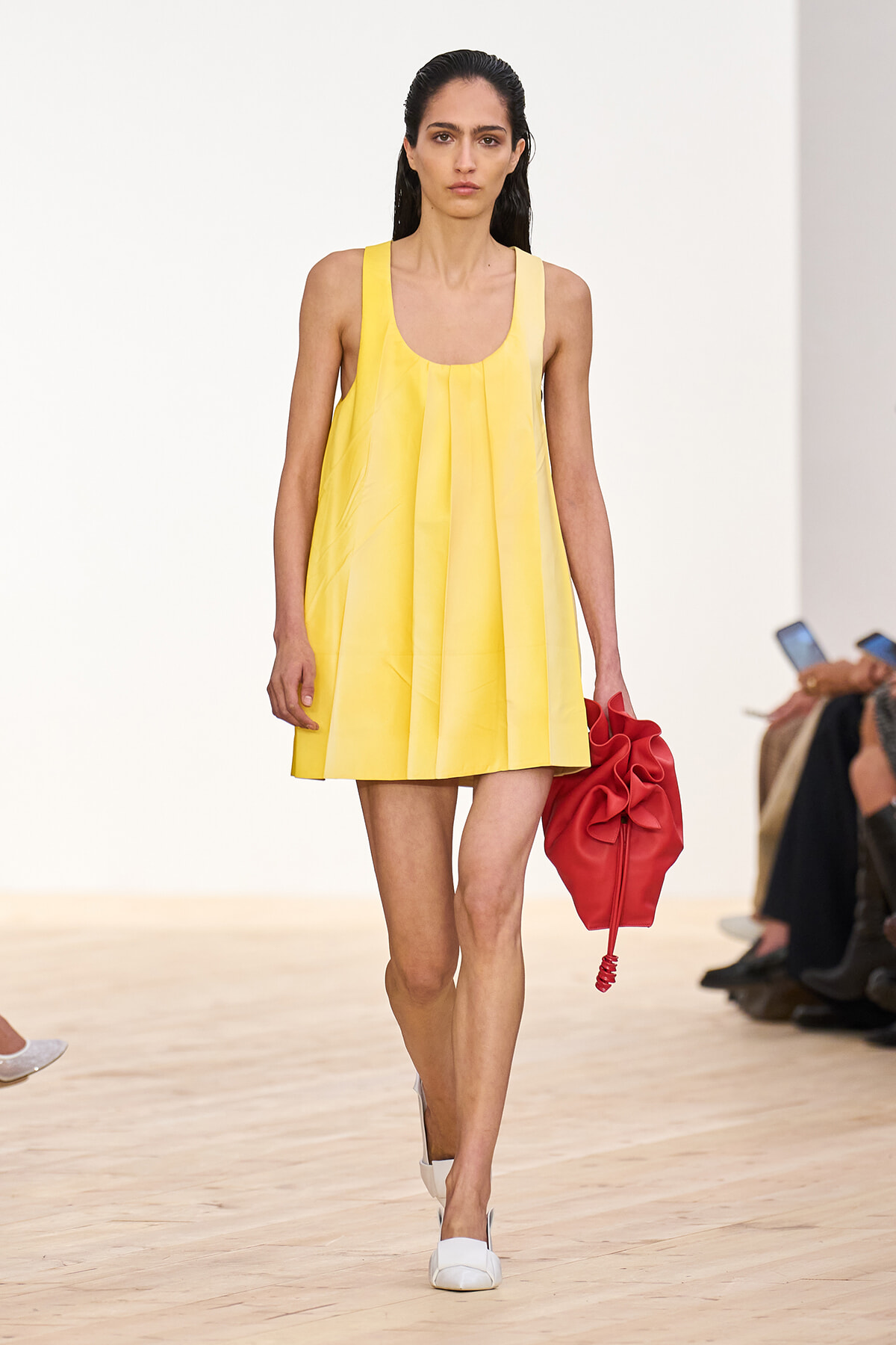 Model walking runway in a bright yellow sleeveless pleated dress, carrying a red handbag and wearing white shoes.