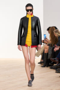 Model walks the runway in a black leather blazer over a bright yellow outfit with red shorts, wearing oversized black sunglasses.
