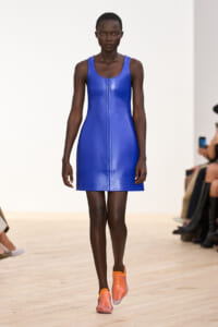 Model walking the runway in a cobalt blue sleeveless zip-front mini dress with orange-pink slip-on shoes.