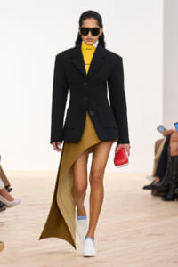 Model walking a runway in a black blazer over a mustard turtleneck and asymmetric skirt, white and blue shoes, and oversized sunglasses.
