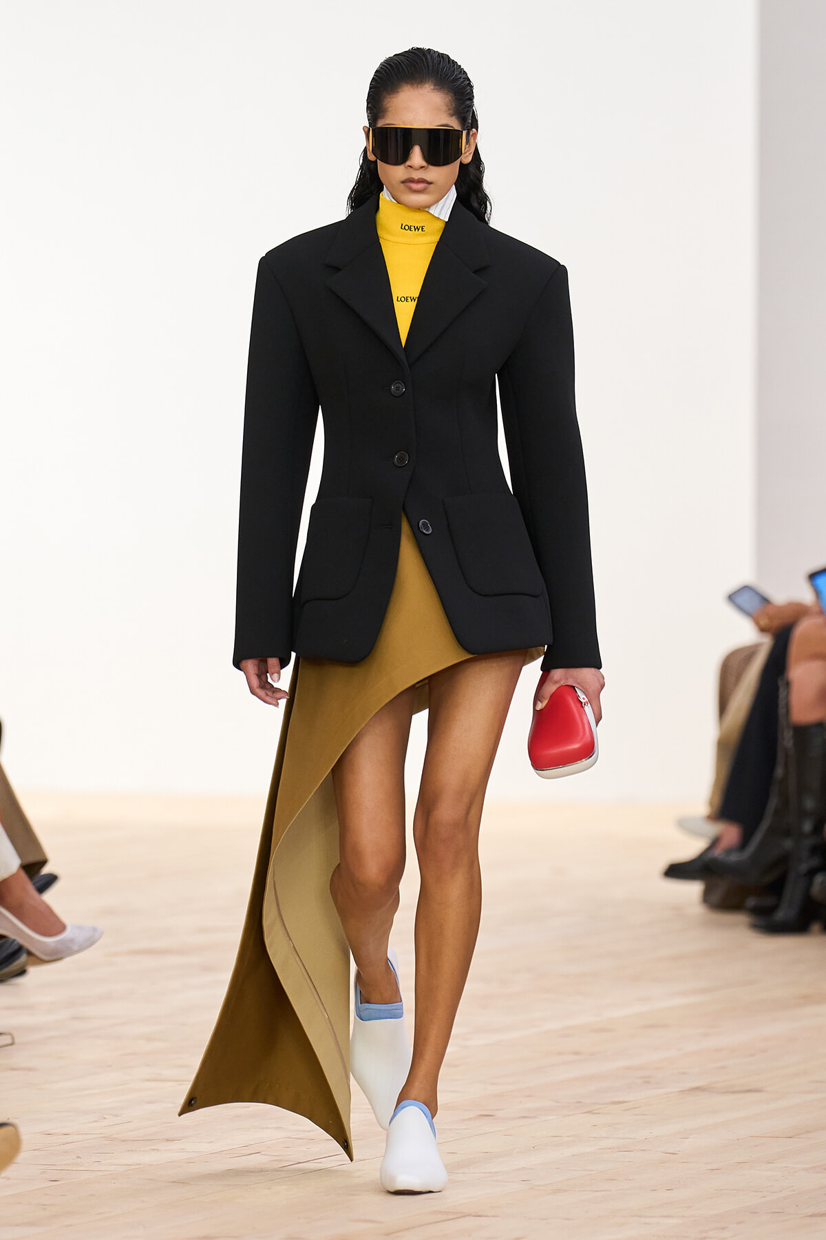 Model walking a runway in a black blazer over a mustard turtleneck and asymmetric skirt, white and blue shoes, and oversized sunglasses.