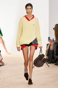 Model walking a fashion show in a cream oversized sweater, red accent top, black shorts with red underlayer, and a brown handbag.