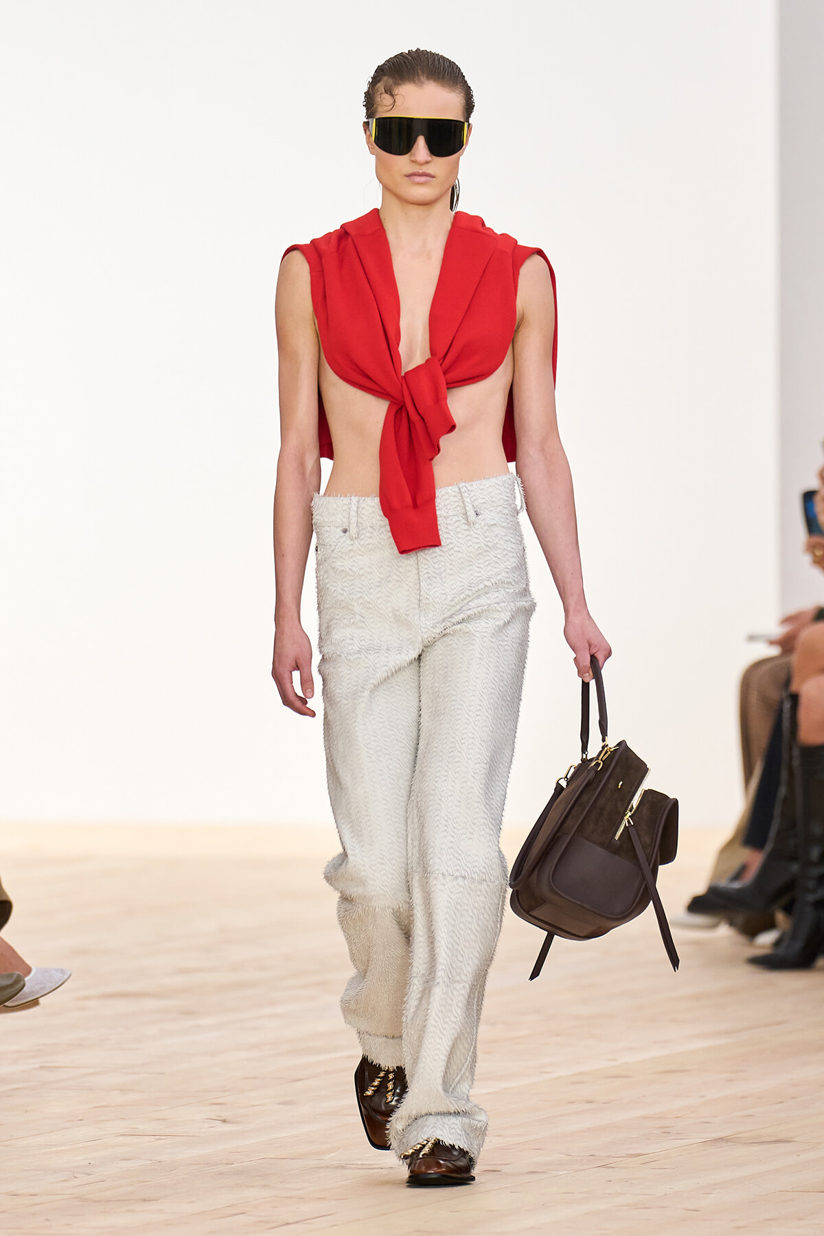 Model walking a runway in a red sleeveless top tied at the front, textured white wide-leg pants, oversized sunglasses, and a brown handbag.