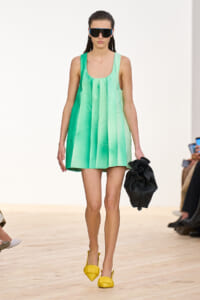 Model on a runway wearing a mint green sleeveless pleated dress, oversized black sunglasses, yellow heels, and a black handheld bag.
