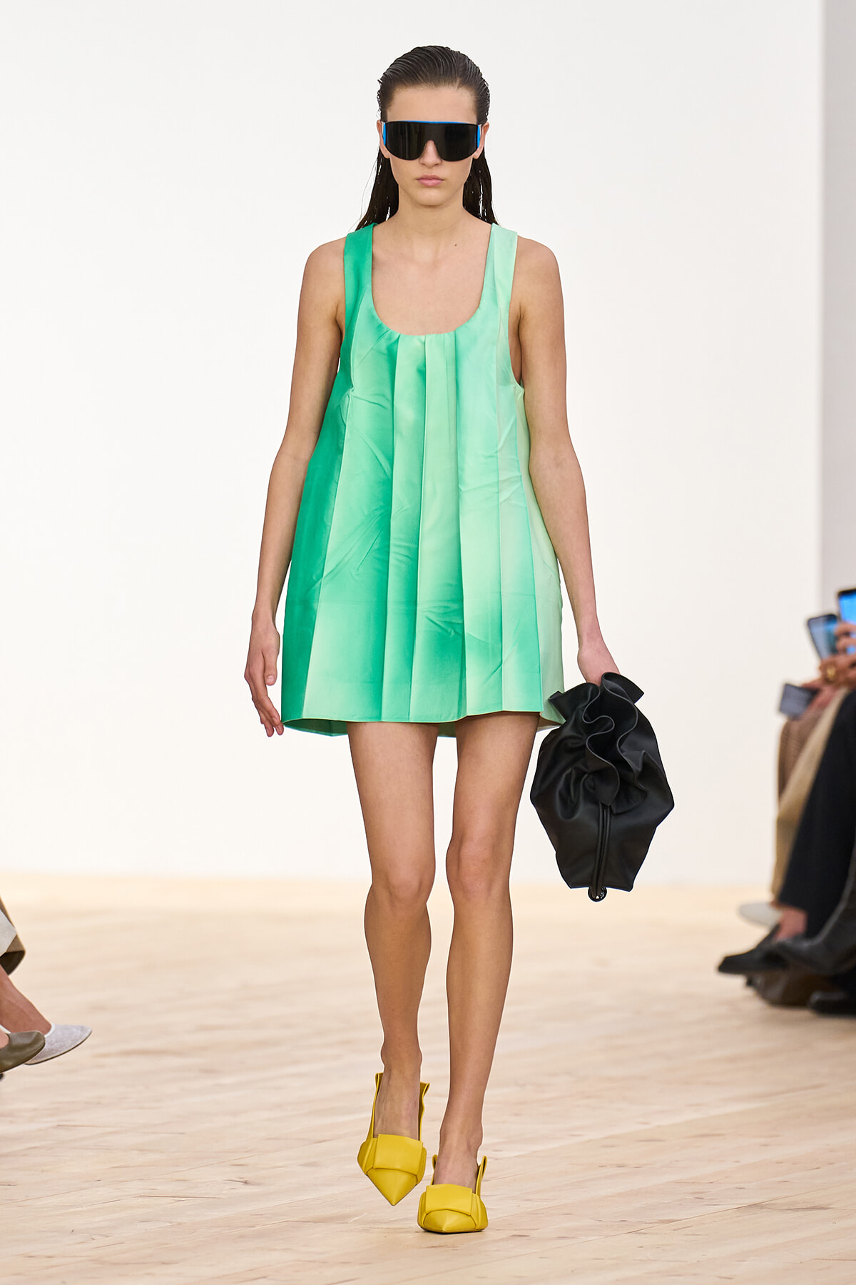 Model on a runway wearing a mint green sleeveless pleated dress, oversized black sunglasses, yellow heels, and a black handheld bag.