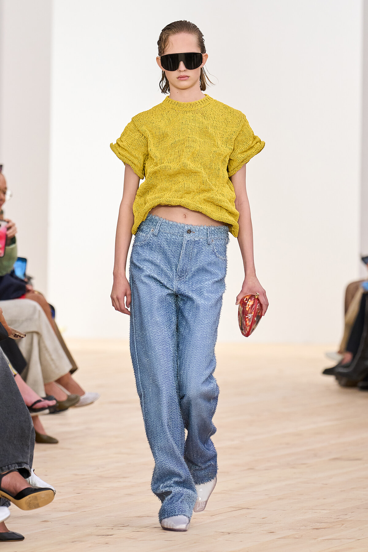 Model walking a fashion runway in a mustard yellow knit top, wide-light denim jeans, oversized black sunglasses, and a small red clutch clutched in her right hand