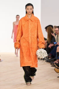 Model on a runway wearing an orange textured coat, carrying a white petal-inspired handbag with fringe.