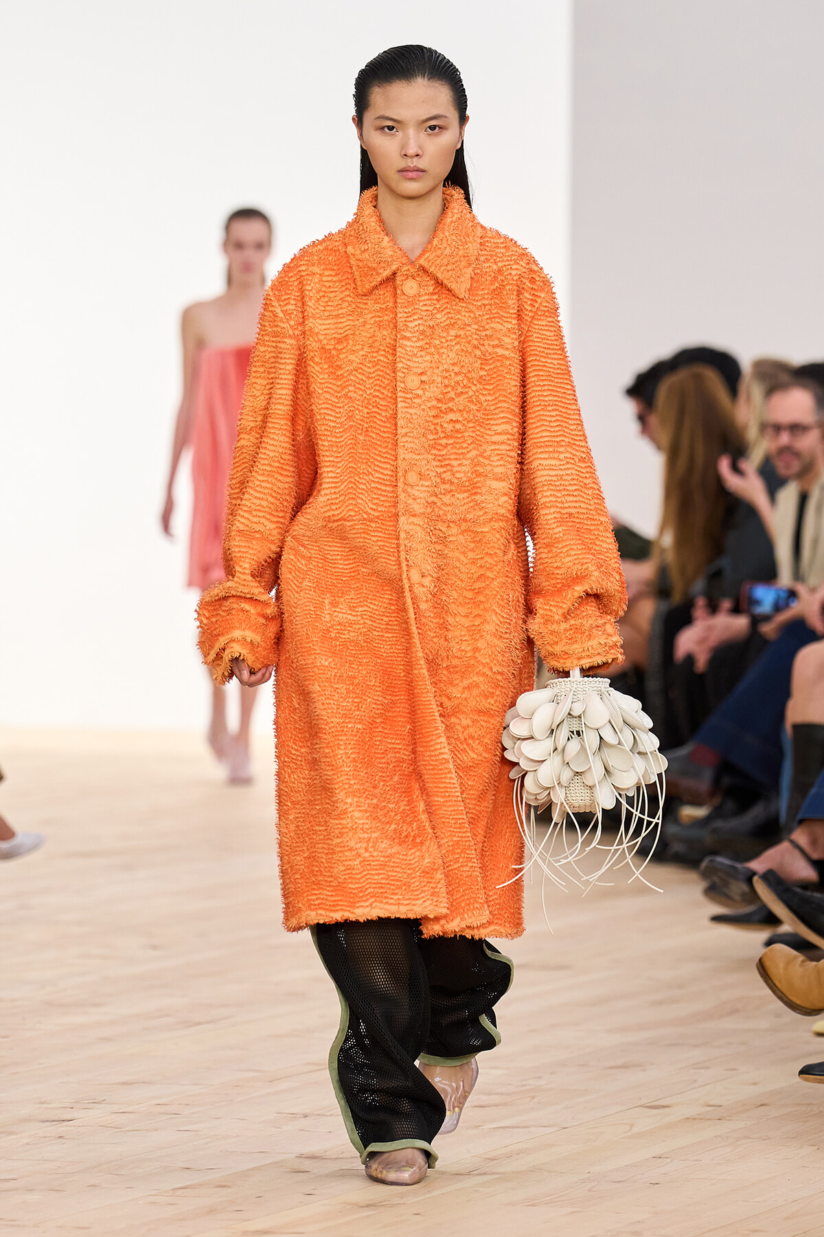 Model on a runway wearing an orange textured coat, carrying a white petal-inspired handbag with fringe.
