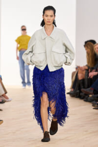 Model walks the runway in an oversized light gray jacket and a royal blue fringe skirt with a high slit, brown shoes.