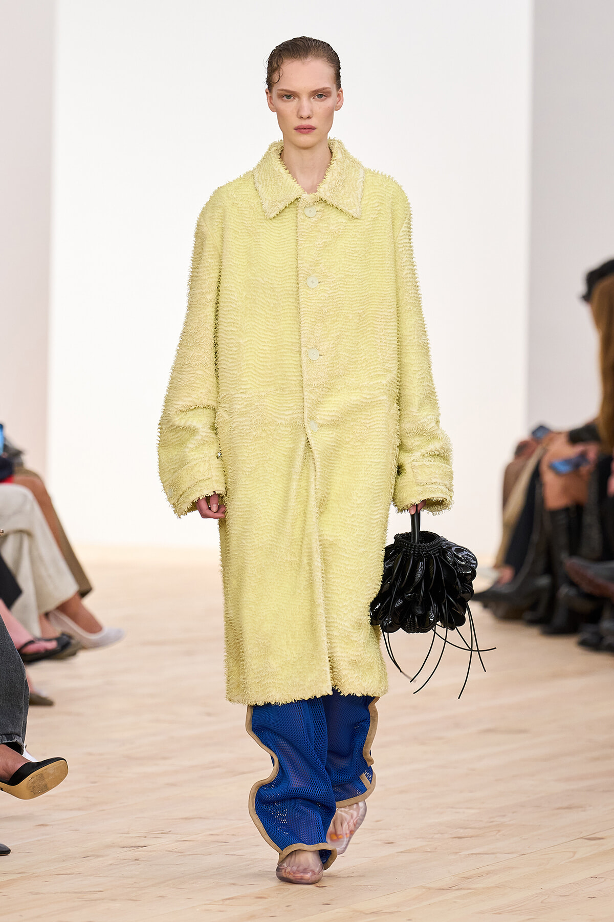 Model on a runway wearing a pale yellow textured oversized coat with blue pants and a black sculptural handbag.