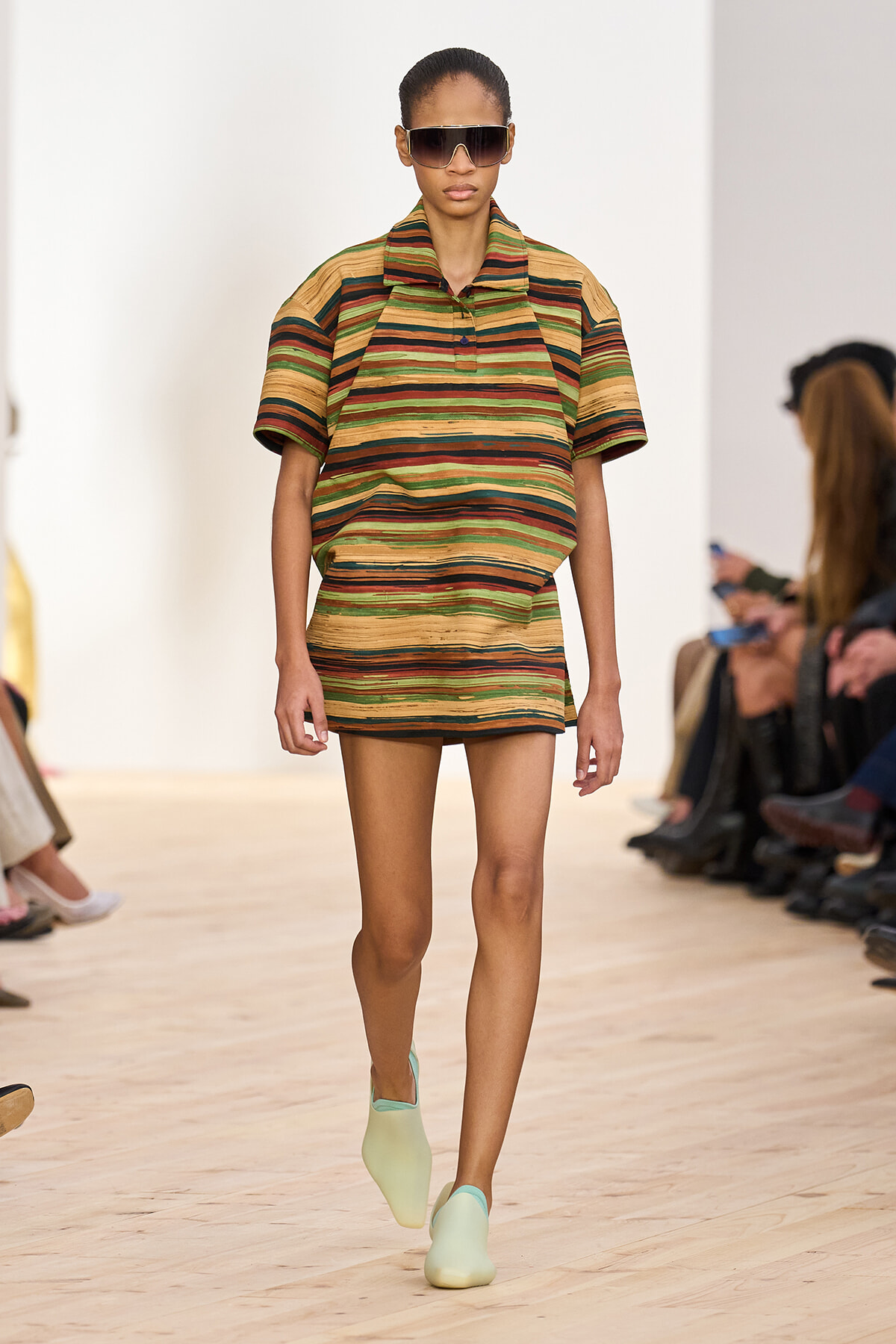 Model walking runway in a green-brown striped, short-sleeve polo dress, wearing oversized sunglasses and mint-green shoes