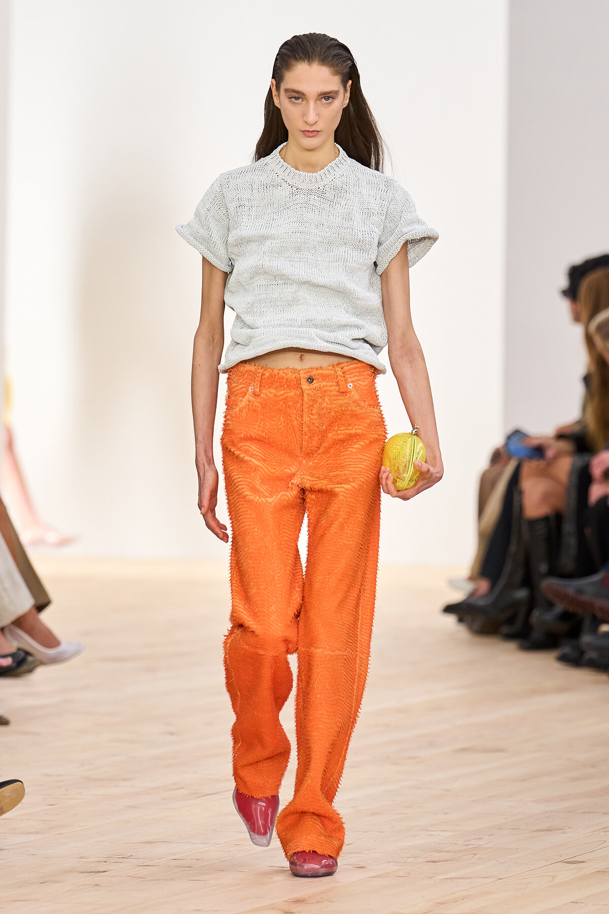 Model walking a runway in a light gray knit top and bright orange textured pants, holding a yellow clutch accessory.