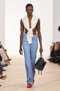 Model walks the runway in a sleeveless cream wrap-top tied at the front, paired with light-wash distressed jeans and bright red shoes, carrying a black handbag.