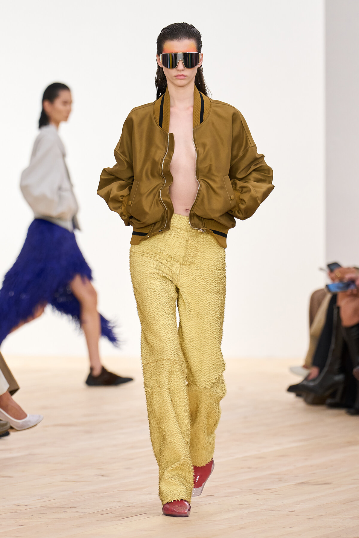 Model walking a runway in an oversized brown bomber jacket, textured yellow pants, red shoes, and futuristic reflective sunglasses.