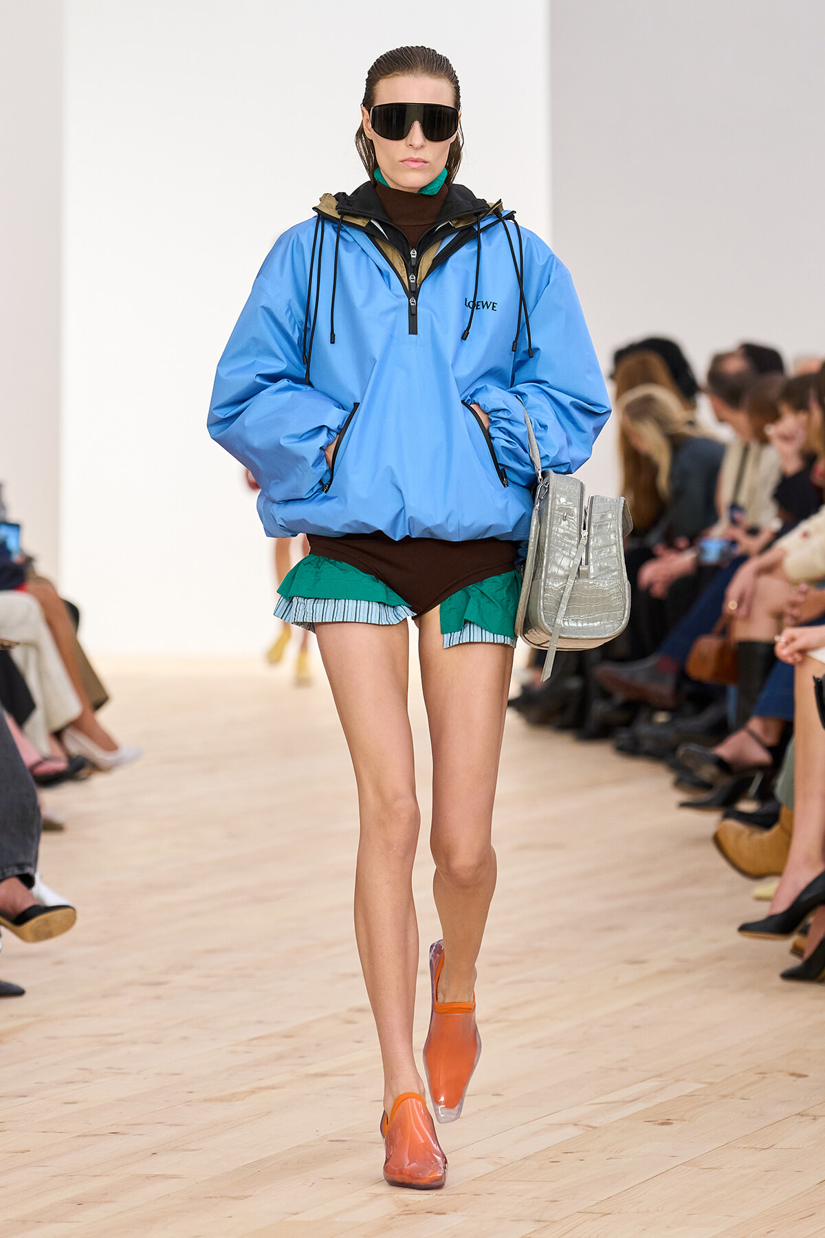 Runway model in a bright blue LOEWE windbreaker, layered teal shorts, orange shoes, and oversized sunglasses walking the catwalk with a silver bag on her shoulder.