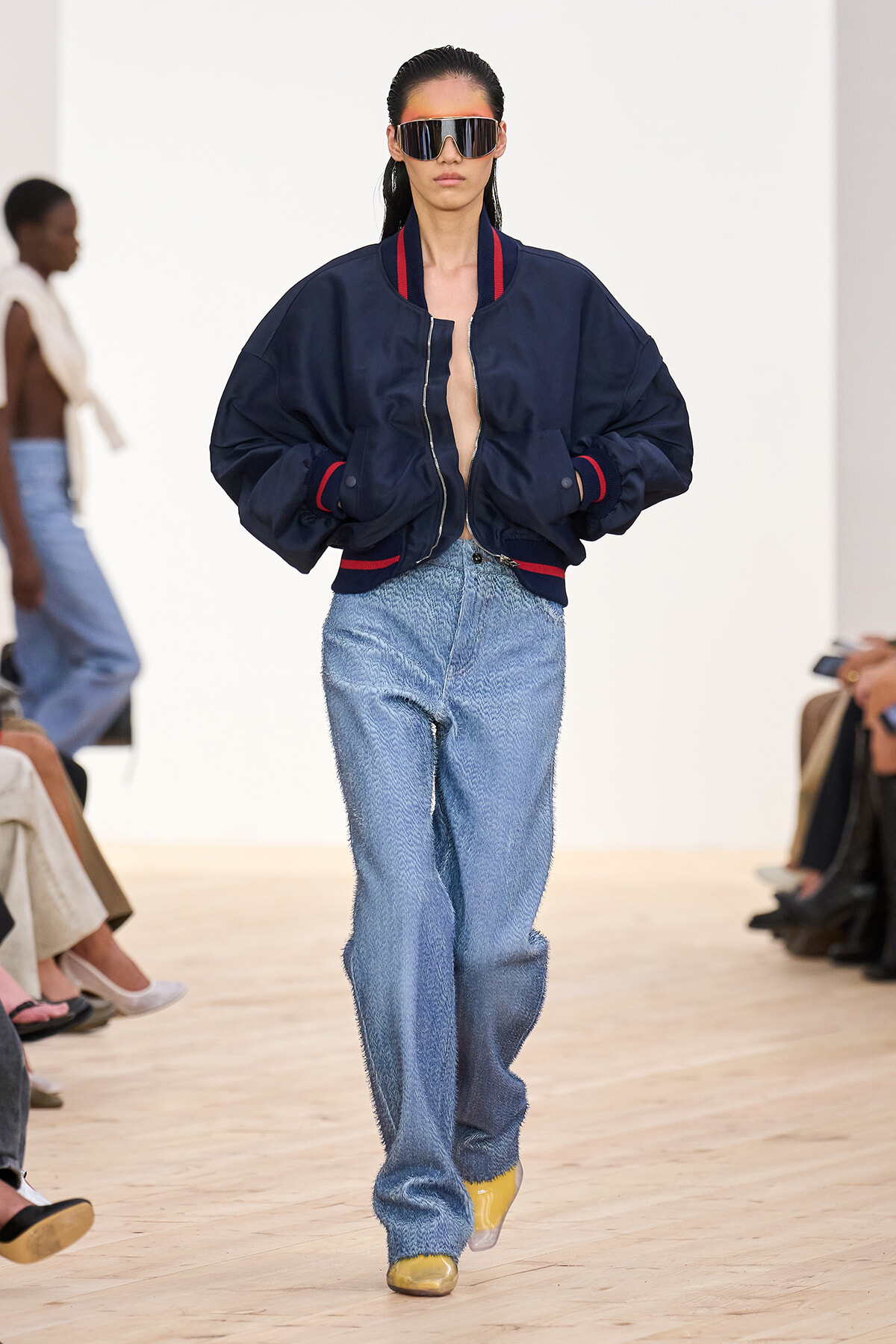 Model walking the runway in an oversized navy bomber jacket with red trim, wide-leg jeans, and yellow shoes, wearing reflective sunglasses.