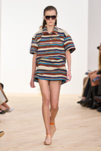 Model walking the runway in a multicolored horizontal striped mini dress with short sleeves and oversized sunglasses.