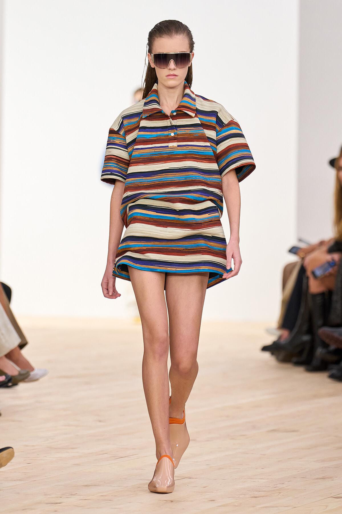 Model walking the runway in a multicolored horizontal striped mini dress with short sleeves and oversized sunglasses.