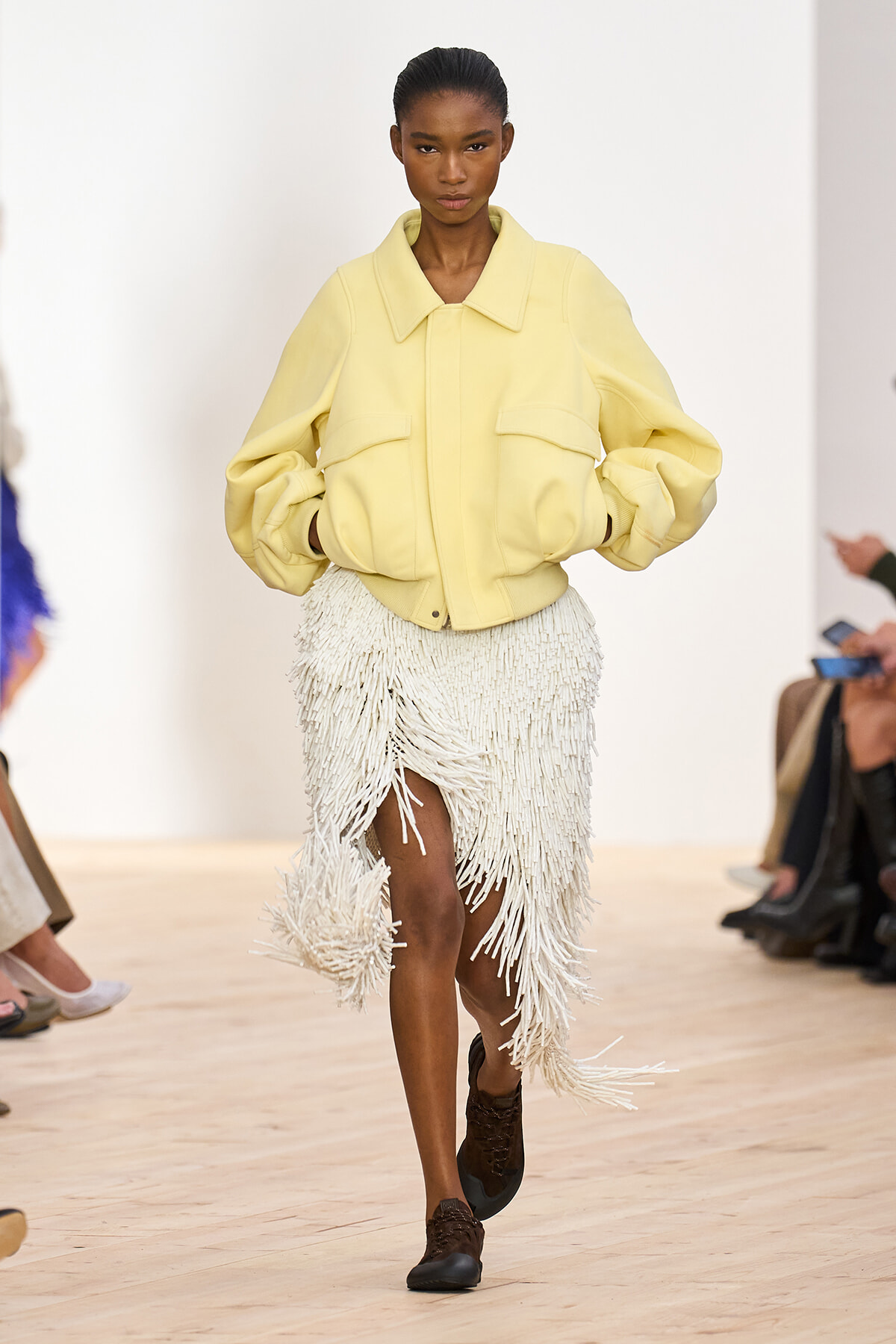 Model walking the runway in a pale yellow oversized jacket with large front pockets, paired with a white fringe skirt and brown lace-up shoes.
