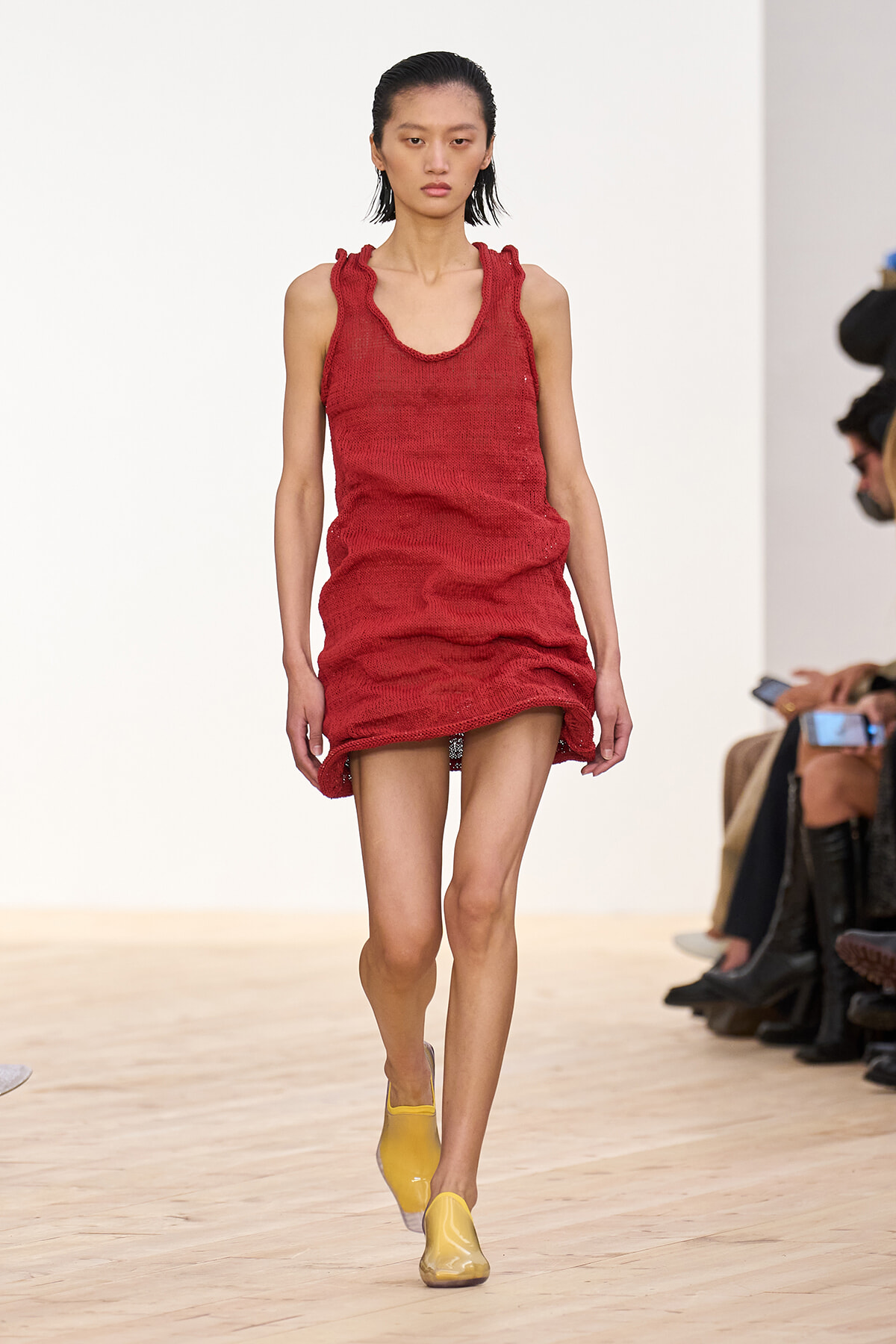 Model walks down a runway in a red knit sleeveless mini dress with gathered neckline and hem, paired with yellow flats.