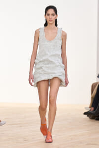 Model walking a runway in a cream, sleeveless knit dress with ruffled straps and a loose, ruched hem; orange clogs complete the look.